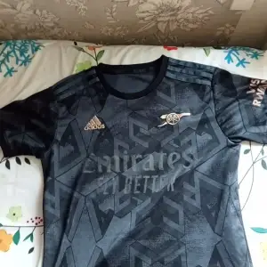 Snygg svart fotbollströja från Adidas med geometriskt mönster och korta ärmar. Tröjan har guldiga detaljer och en logotyp på bröstet. Perfekt för fotbollsfans som vill ha en stilren look.
