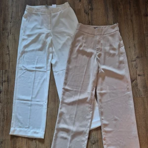 Två par kostymbyxor i beige och vit - Säljer två par eleganta kostymbyxor i beige och vit. Byxorna har en högmidjad design med en rak passform som ger en stilren look. Perfekta för en chic och sofistikerad stil.