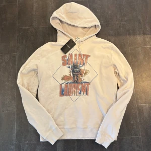 Saint Laurent Hoodie - Tja! Säljer denna riktigt feta beige/vita Saint Laurent hoodien i nyskick! Hoodien är somsagt i nyskick och har inga tecken på använding alls. Slitningarna på luvan och muddarna är design. Nypris ca 9000kr. Size S men stor i storleken så skulle säga M. Självklart äkta. Skriv vid funderingar!