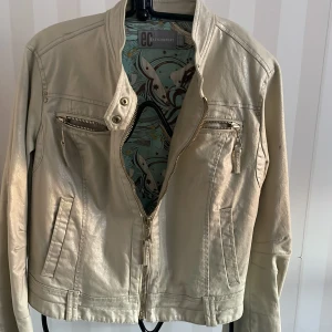 Beige jacka från East  Comfort st 42/44 - Snygg beige med guldbrons glitter jacka från EC Envy Comfort med dragkedja och flera praktiska fickor. Jackan har en stilren design med långa ärmar och en unik mönstrad insida. Perfekt för en trendig look!