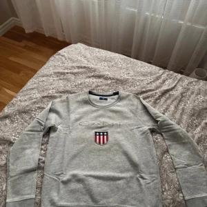Grå sweatshirt från Gant - Säljer en stilren grå sweatshirt från Gant med broderad logga och ett amerikanskt sköldmärke på bröstet. Tröjan har långa ärmar och rund halsringning. Perfekt för en avslappnad stil.