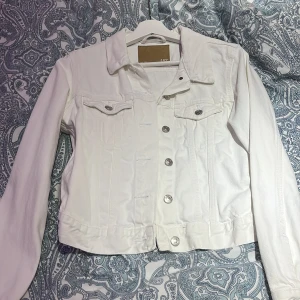 Vit jeansjacka från 157 - Säljer en stilren vit jeansjacka från 157 i storlek XS. Jackan har långa ärmar och knäpps med silverfärgade knappar framtill. Två bröstfickor med knappstängning ger en klassisk look. Perfekt för vår och sommar!