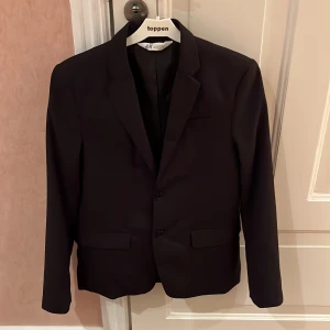 Svart kavaj från H&M - Snygg svart kavaj från H&M i storlek 164. Kavajen har en klassisk design med två knappar framtill och fickor med lock. Perfekt för formella tillfällen eller som en stilren vardagskavaj.