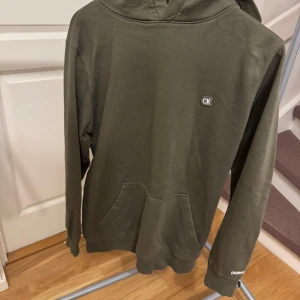 Olivgrön hoodie från Calvin Klein - Säljer en stilren olivgrön hoodie från Calvin Klein. Tröjan har en klassisk passform med långa ärmar och en praktisk magficka. CK-logga på bröstet och Calvin Klein Jeans-tryck på ärmen ger en snygg touch. Storlek: S. Nypris: 1200
