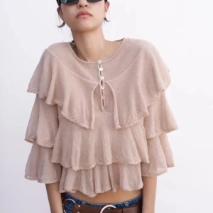 Beige volangblus med knappar - Säljer en chic beige blus med flera lager av volanger och knappar framtill. Blusen har en lös passform och trekvartsärmar, perfekt för en stilfull look.