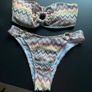 Mönstrad bikini med ringdetaljer - Trendig bikini med ett färgglatt, vågigt mönster i rosa, gul, grön och svart. Överdelen har en unik ringdetalj i mitten och underdelen har matchande ringar på sidorna. Perfekt för en dag på stranden!