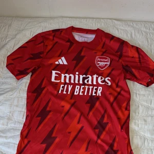 Röd Arsenal fotbollströja - Säljer en röd Arsenal fotbollströja från Adidas med ett blixtmönster i mörkrött. Tröjan har korta ärmar och vit text med sponsorlogga på framsidan. Perfekt för alla Arsenal-fans! ⚽️