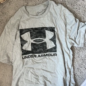 Grå t-shirt från Under Armour - Säljer en grå t-shirt från Under Armour med ett stort logotryck i svart och camouflage på framsidan. T-shirten har en lös passform och är kortärmad, perfekt för en avslappnad stil.