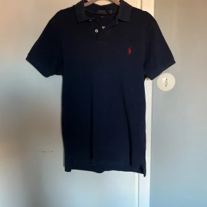 Mörkblå pikétröja från Ralph Lauren - Säljer en stilren mörkblå pikétröja från Ralph Lauren i slim fit. Tröjan har korta ärmar och en klassisk krage med knappar. Den ikoniska loggan är broderad i rött på bröstet. Perfekt för en avslappnad men ändå elegant look.
