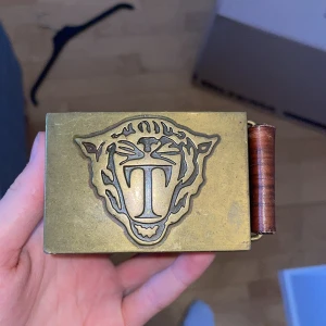 Brunt läderbälte från Tiger of Sweden ( - Snyggt brunt läderbälte från Tiger of Sweden med en unik mässingsfärgad spänne som har ett tigerhuvud och bokstaven T. Perfekt för att ge en stilren touch till din outfit.