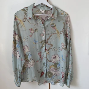 Dimgrön vårblus med fågelmotiv från GP&J BAKER - Vårlig blus i dimgrönt med ett mönster med fåglar, fjärilar och blommor i mjuka pasteller. Tunn och böljande. Pärlemorknappar. GP&J BAKER för H&M. Längd 70 cm, mått runt midjan 108 cm. Storlek 42. 