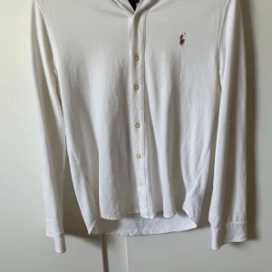 Vit skjorta från Ralph Lauren - Säljer en stilren vit långärmad skjorta från Ralph Lauren med klassisk krage och knappar framtill. Skjortan har en broderad logotyp på bröstet som ger en elegant touch. Perfekt för en snygg och avslappnad look. Den har även ett luftigare material som är perfekt för sommaren