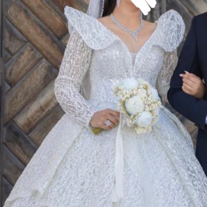 Vit glittrig brudklänning - Elegant vit brudklänning med glittrande detaljer och lång ärm. Klänningen har en v-ringad design med volanger vid axlarna och en imponerande lång kjol. Perfekt för en sagolik bröllopsdag.