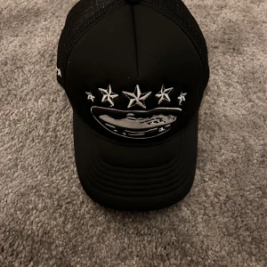 Corteiz keps - Snygg svart truckerkeps från Corteiz med broderade stjärnor och ett motiv framtill. Kepsen har ett nätmaterial på sidorna och justerbar passform. Perfekt för en cool och avslappnad stil.