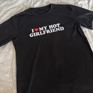 Svart t-shirt med tryck - Säljer en svart t-shirt med texten 'I ❤️ MY HOT GIRLFRIEND' i vitt och rött. Perfekt för att visa din lojalitet till din tjej. Tyvärr så blev mitt ex konsumerad av gatan så nu måste jag bli av med den 🤷🏽‍♂️