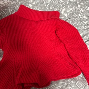 Röd stickad polotröja - Säljer en snygg röd stickad polotröja med ribbad design. Perfekt för kyliga dagar med sin höga krage som går snett till armen och långa ärmar. Tröjan har en härlig struktur och är både stilren och bekväm.