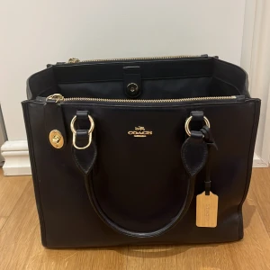 Marinblå handväska från Coach  - Snygg marinblå handväska från Coach i skinn med guldfärgade detaljer köpt från NK. Väskan har både handtag och en avtagbar axelrem, samt dragkedja för säker förvaring. Perfekt för att bära med sig det viktigaste med stil. Inga skador, inga fel precis som ny ✨