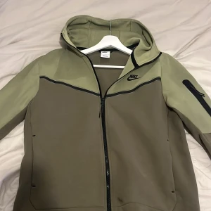 Grön Nike Tech - Säljer en snygg grön hoodie från Nike med dragkedja och huva. Jackan har långa ärmar och en praktisk ficka med dragkedja på ärmen. Perfekt för en skön look. Kan sänka pris vid snabb affär