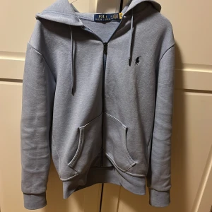 Ljusblå hoodie från Ralph Lauren - Säljer en stilren ljusblå hoodie från Ralph Lauren med dragkedja och justerbar huva. Tröjan har långa ärmar och en liten broderad logga på bröstet. Perfekt för en avslappnad stil. 🩵