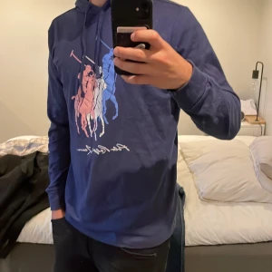 Blå hoodie Polo Ralph Lauren M - Snygg blå hoodie från Polo Ralph Lauren i bomull. Framsidan har ett stort tryck med polospelare i blått, vitt och rött. Tröjan har huva med snörning och långa ärmar. Perfekt för dig som gillar streetstyle och klassiska märken.