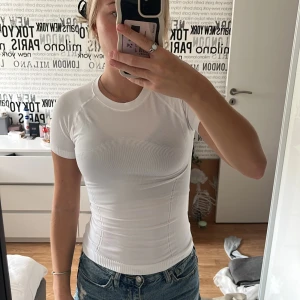 Träningstopp - Säljer en vit, tight ribbad träningstopp med korta ärmar och rund hals. Toppen har en figurnära passform och är tillverkad i ett stretchigt material som sitter snyggt på kroppen. Aldrig använd!