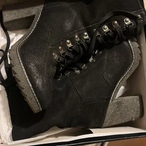 Svarta kängor med grov sula - Tuffa svarta kängor i skinn med grov, grå sula och snörning med metallhakar. Perfekta för dig som vill ha en cool och robust look. Kängorna har rund tå och markerad klack för extra attityd.