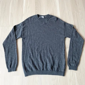 Mörkgrå stickad tröja i ullmix - En stilren mörkgrå stickad tröja med rund hals och långa ärmar. Tröjan är tunn och mjuk, perfekt för lager-på-lager. Ribbstickade muddar vid ärmslut och nederkant ger en snygg passform. Passar dig som gillar en enkel och clean look.