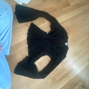Svart omlottopp med volangärm - Säljer en svart omlottopp med knyt i midjan och långa, vida volangärmar. Toppen är ribbad och har en lätt transparent känsla med snygg struktur. Perfekt för dig som gillar trendiga detaljer och vill sticka ut lite extra. Fråga om mer bättre bilder Aldrig använd 🥰