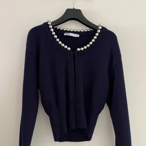 Mörkblå kofta med pärldetaljer från Zara - Snygg mörkblå kofta från Zara med stora pärldetaljer runt halsringningen. Modellen är croppad med ribbad nederkant och långa ärmar. Perfekt för dig som gillar att sticka ut med coola detaljer och vill ha en unik look.