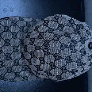Gucci keps med GG-mönster - Snygg keps från Gucci med klassiskt GG-mönster i svart på beige bakgrund. Kepsen har en böjd skärm och en detalj med röd och marinblå rand baktill. Tillverkad i bomull och polyester, insidan är fodrad i mörkblått tyg. Perfekt för dig som gillar lyxiga accessoarer.
