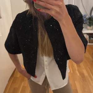 Svart paljettjacka från Maje - Säljer en svart, croppad cardigan jacka från Maje med paljettdetaljer. Jackan har en öppen front, kortare passform och är tillverkad i ett grovt, stickat material som ger en lyxig känsla. Perfekt för dig som vill sticka ut med stilrena detaljer.