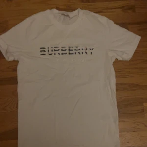 Vit burberry t shirt - Snygg vit t-shirt från Burberry med logotypen tryckt i svart och grått på bröstet. Perfekt för en stilren och klassisk look.