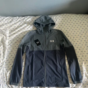 Grå vindjacka från Under Armour - Snygg tvåfärgad vindjacka från Under Armour i mörkgrått och ljusgrått. Jackan har huva, dragkedja framtill och logga på bröstet. Elastiska muddar med tryckt text på ärmsluten. Perfekt för aktiva dagar utomhus. Jackan är använd ett fåtal gånger. Vid fler bilder eller frågor är det bara att skriva.