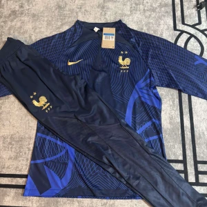 Frankrike Nike fotbollströja blå/guld - Säljer en officiell Frankrike fotbollströja från Nike i mörkblått med guldiga detaljer och FFF-logga. Tröjan har lång ärm, halv dragkedja och ett coolt geometriskt mönster i blå toner. Materialet är lätt och andas, perfekt för träning eller match.