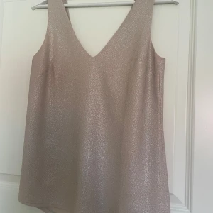 Glittrig champagnefärgad linnetopp Oasis - Supersnygg linnetopp från Oasis i champagnefärg med glittrig finish. Toppen har v-ringning både fram och bak och är ärmlös med breda axelband. Perfekt för dig som vill sticka ut lite extra med en stilren men ändå festlig look.