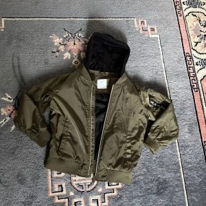 Olivgrön bomberjacka från Lindex Kids - Säljer en olivgrön bomberjacka från Lindex Kids med svart huva och cool detalj med texten 'URB_N W_RLD' på ärmen. Jackan är vattenavvisande och har dragkedja framtill samt ribbade muddar vid ärmslut och midja. Perfekt för dig som gillar streetstyle. Priset kan diskuteras 