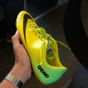 Nike Mercurial gula fotbollsskor - Säljer ett par Nike Mercurial fotbollsskor i en riktigt fet gul färg med svarta detaljer och gröna inslag. Skorna har snörning, platt sula med dobbar och är gjorda i syntetmaterial. Perfekta för dig som vill sticka ut på planen och få grymt grepp.