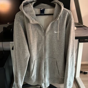 Grå hoodie med dragkedja från Nike - Snygg grå hoodie från Nike med dragkedja och huva. Tröjan har vita snören, två fickor framtill och Nike-logga på bröstet. Mjuk insida och klassisk passform, perfekt för chill eller träning. Enkel och stilren design som funkar året runt.