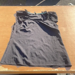 Svart ärmlös topp från Vero Moda - Svart ärmlös topp från Vero Moda med lätt puffade axlar och dekorativ snodd vid halsringningen. Toppens material känns mjukt och följsamt, perfekt för att styla med jeans eller kjol. Enkel och stilren design som passar till många olika looks.