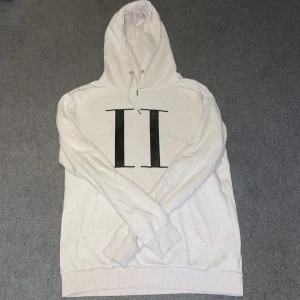 Vit hoodie från Les Deux med tryck - Snygg vit hoodie från Les Deux med stort grön-svart II-tryck på bröstet. Klassisk modell med huva, samt känguruficka framtill. Mjuk bomullsblandning som är perfekt för chill dagar eller när du vill ha en clean look.
