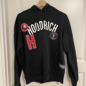 Svart hoodie från Hoodrich med tryck - Svart hoodie från Hoodrich med stora vita och röda tryck och patches framtill. Klassisk huva med dragsko och känguruficka. Hoodrich-logga på bröstet och ärmen. Perfekt för dig som gillar streetwear och vill sticka ut.