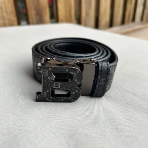 Svart burburry bälte med B-spänne - Snyggt svart bälte från Balmain med ett stort, glansigt B-format metallspänne. Bältet har diskret mönster i grått och svart längs hela remmen och är gjort i skinn. Perfekt accessoar för att lyfta din outfit. Äkthet garanteras ej. Därav priset!