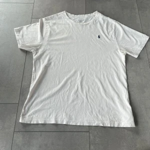 Vit t-shirt från Polo Ralph Lauren - Klassisk och stilren vit T-shirt från Ralph Lauren. Tidlös design med den ikoniska loggan på bröstet. T-shirten är i bra skick, lätt använd. Perfekt för både vardag och fritid! 	•	Storlek: S (Barn XL) – passar ungefär som en liten vuxenstorlek 	•	Färg: Vit 	•	Skick: Bra – inga hål eller stora fläckar 	•	Mjukt och bekvämt material