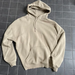 Beige hoodie med huva och ficka - En stilren beige hoodie med huva och dragsko, samt en stor magficka framtill. Tröjan har långa ärmar och ribbade muddar vid ärmslut och nederkant. Märken på huven som inte lossnar vid tvätt.