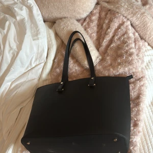 Svart axelväska i skinn med dragkedja - Stilren svart axelväska i skinn med raka linjer och långa handtag. Väskan har en praktisk dragkedja i guld och metallfötter undertill. Perfekt storlek för att få plats med allt du behöver till skolan eller stan.