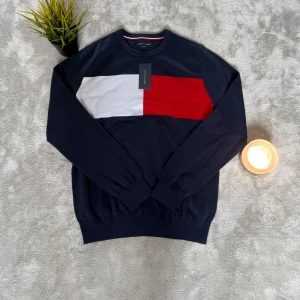 Mörkblå Tommy Hilfiger stickad tröja - Snygg mörkblå stickad tröja från Tommy Hilfiger med breda block i vitt och rött över bröstet. Klassisk rund halsringning med diskret logga på ärmen och ribbade muddar. Perfekt för dig som gillar stilren design med en sportig touch.