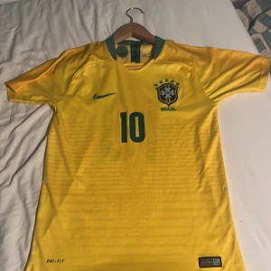 Brasilien Neymar Jr 10 Nike fotbollströja - Gul Brasilien fotbollströja från Nike med gröna detaljer, Neymar Jr och nummer 10 på ryggen. Klassisk landslagströja med Dri-FIT-material som andas och korta ärmar. Perfekt för dig som vill köra stenhårt på planen eller visa support för Brasilien. Från år 2018