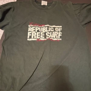 Svart Gildan Free Surf t-shirt L - Cool svart t-shirt från Gildan med trycket 'Republic of Free Surf' i vitt och rött på bröstet och mindre tryck på ryggen. Klassisk passform med rund hals och korta ärmar. Tillverkad i 100% bomull för skön känsla och bra kvalitet.