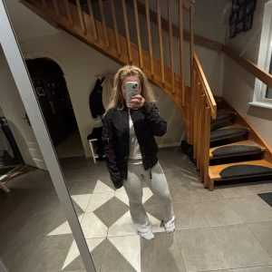 Svart bomberjacka från Alpha Industries - Säljer en svart bomberjacka från Alpha Industries med klassisk orange insida och ikonisk 'Remove Before Flight'-tag på ärmen. Jackan har dragkedja, ribbade muddar och lång ärm. Perfekt för dig som vill ha en streetig och tidlös look.