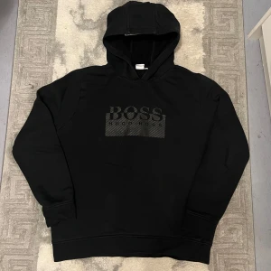 Svart hoodie från Hugo Boss - Svart hoodie från Hugo Boss är en gammal rare model med stor logga på bröstet i ton-i-ton. Klassisk huva, långa ärmar och mjukt material som känns skönt mot huden. Perfekt för dig som gillar stilrena och snygga streetwear-plagg.